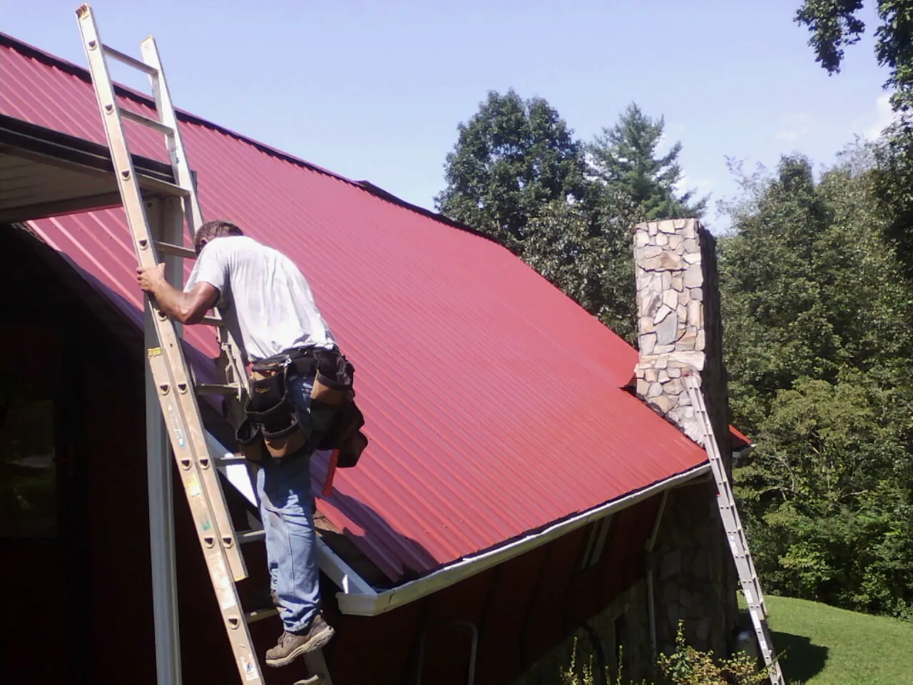 Roofer on a ladder beside a red metal roof and stone chimney — Soffit & Fascia Repair by Roofing Brothers in Mingo